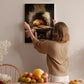 A woman hangs a framed painting of a pizza oven on a wall in a cozy kitchen setting.