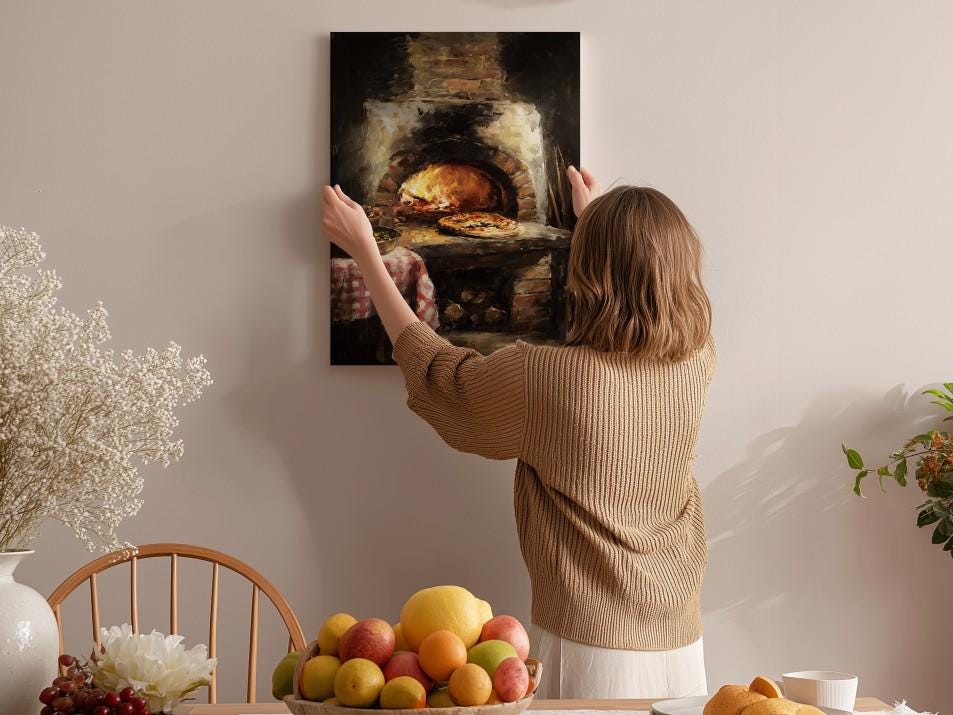 A woman hangs a framed painting of a pizza oven on a wall in a cozy kitchen setting.