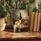 a still life painting of a table with a bottle of wine, cheese, and grapes, surrounded by potted plants and books.
