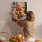 A woman hangs a framed picture of colorful macarons on a wall in a cozy kitchen setting.