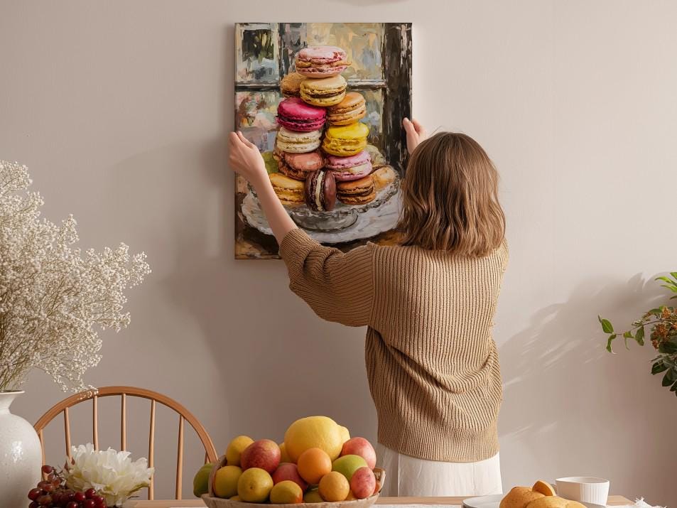A woman hangs a framed picture of colorful macarons on a wall in a cozy kitchen setting.