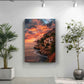 a large framed picture of a coastal cliff with a sunset over the ocean, hanging on a white wall. To the left of the picture, there is a potted plant, and to the right, there is another potted plant.