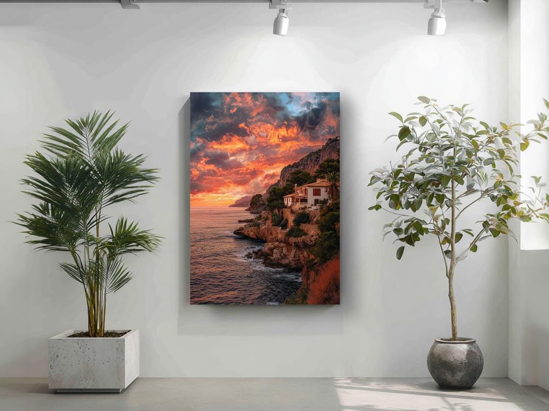 a large framed picture of a coastal cliff with a sunset over the ocean, hanging on a white wall. To the left of the picture, there is a potted plant, and to the right, there is another potted plant.