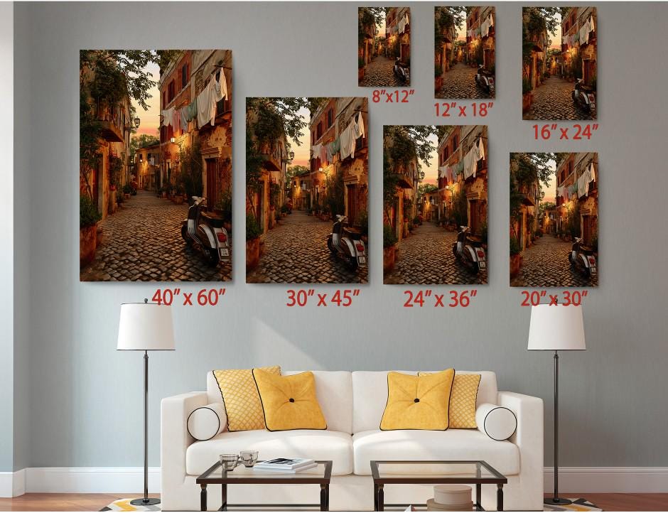 The image is a living room with a white sofa and a coffee table. Above the sofa, there are seven framed photographs of a cobblestone street in different sizes, ranging from 6&#39;x12&quot; to 24&#39;x36&quot;. The photographs are arranged in a grid-like pattern on the wall, with each frame corresponding to a specific size.