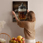 A woman hangs a framed photograph of a glass mug filled with beer on a wall in a cozy kitchen setting.