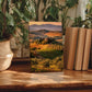 a framed landscape photograph of a countryside scene, placed on a wooden surface next to some potted plants and books.