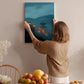 A woman hangs a painting of a serene landscape on a wall, surrounded by a cozy indoor setting with a dining table, chairs, and various fruits.