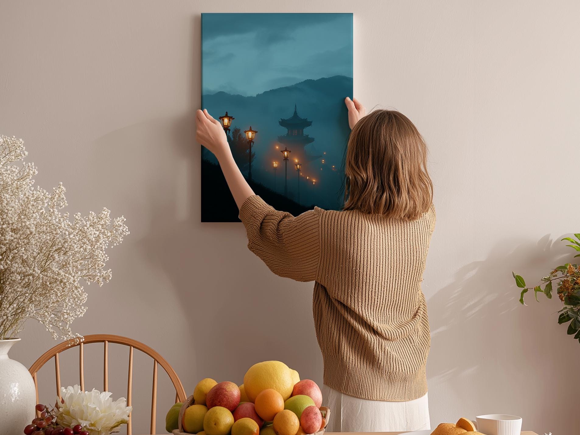 A woman hangs a painting of a serene landscape on a wall, surrounded by a cozy indoor setting with a dining table, chairs, and various fruits.
