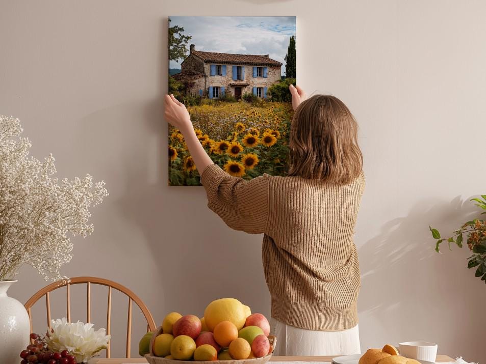 A woman hangs a framed picture of a house with a sunflower field in the background on a wall in a cozy, homely setting.