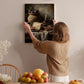 A woman hangs a framed painting of a still life of food and wine on a wall in a cozy, homely setting.