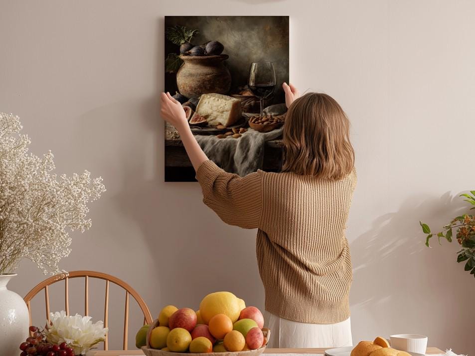 A woman hangs a framed painting of a still life of food and wine on a wall in a cozy, homely setting.