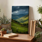 a wooden desk with a framed landscape painting depicting a pastoral scene with sheep grazing in a valley, surrounded by rolling hills and mountains. The painting is placed on a shelf, with several potted plants and books nearby, creating a cozy and natural atmosphere.