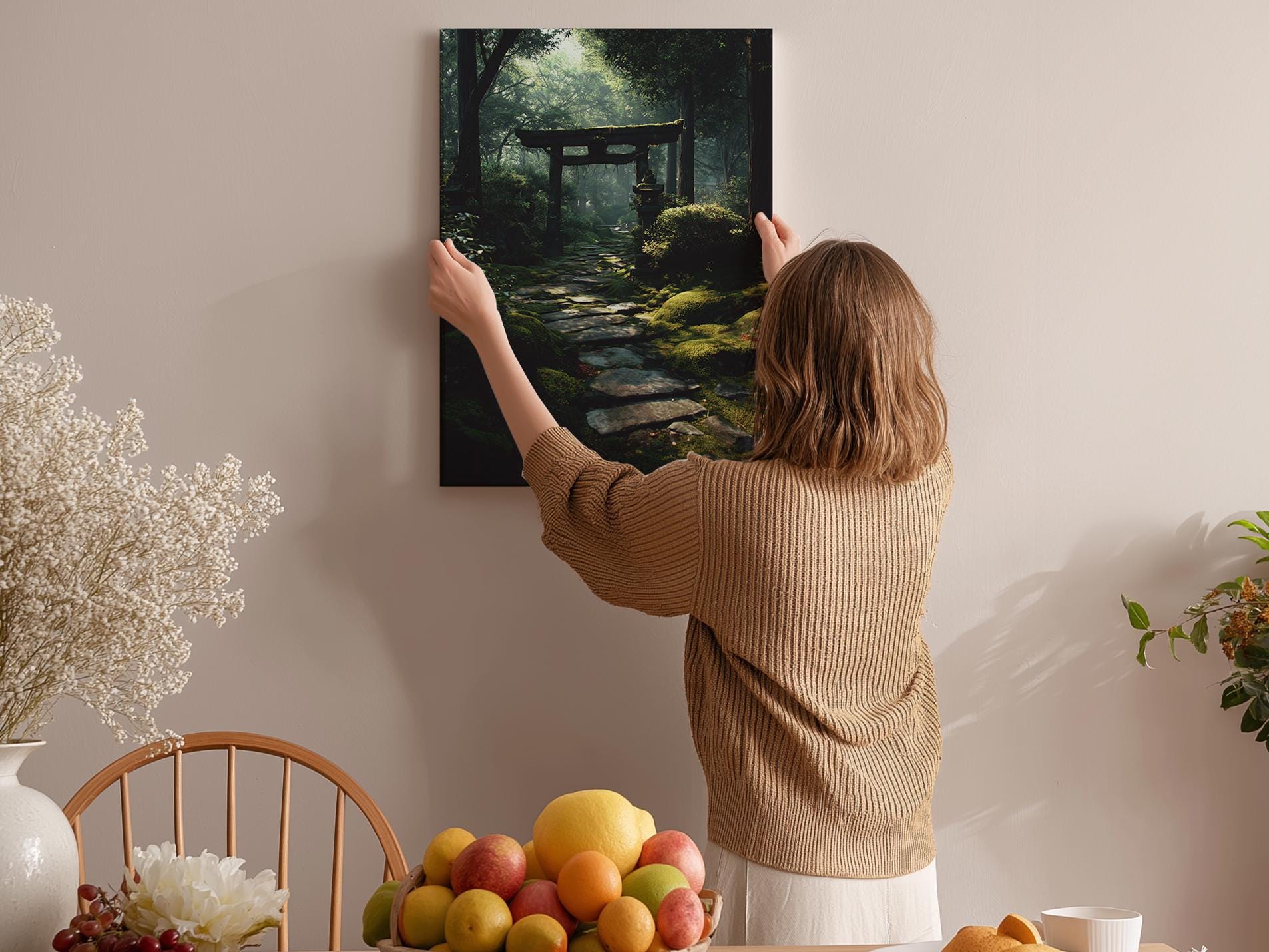 A woman hangs a framed painting of a forest path on a wall in a cozy room with a dining table, chairs, and various fruits.