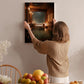 A woman hangs a framed picture on a wall, with a wooden chair, a bowl of fruit, and a vase of flowers in the background.