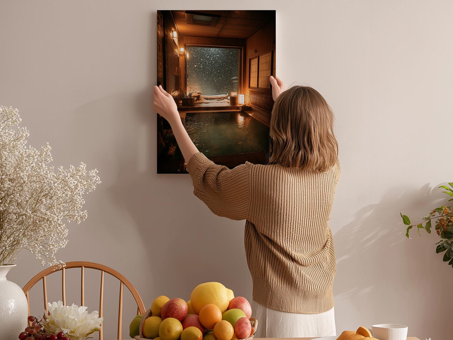 A woman hangs a framed picture on a wall, with a wooden chair, a bowl of fruit, and a vase of flowers in the background.