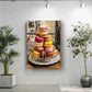 a large painting of a plate of colorful macarons hanging on a wall, with a potted plant to the left and a plant to the right.