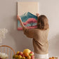 A woman hangs a framed picture of a mountain with a pink tree on a wall in a cozy room with a dining table, chairs, and various fruits.