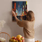 A woman hangs a vibrant painting of a cityscape on a wall in a cozy room, surrounded by a dining table, chairs, and various fruits.