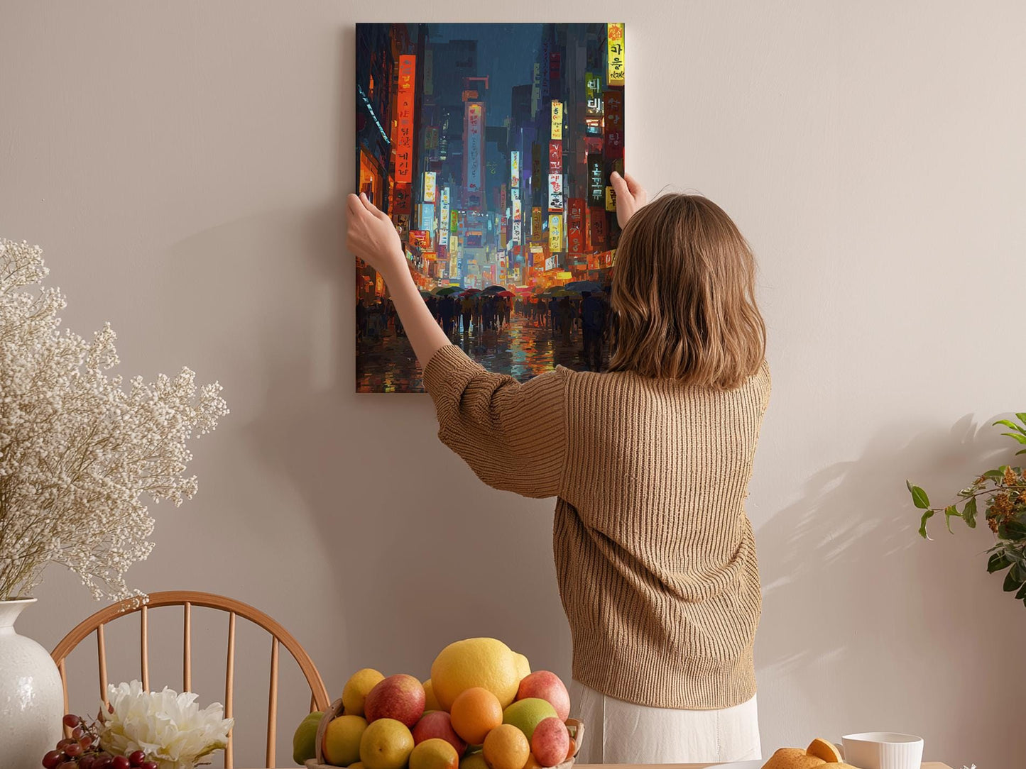 A woman hangs a vibrant painting of a cityscape on a wall in a cozy room, surrounded by a dining table, chairs, and various fruits.