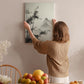 A woman hangs a black and white landscape painting on a wall in a room with a dining table, chairs, and various fruits.