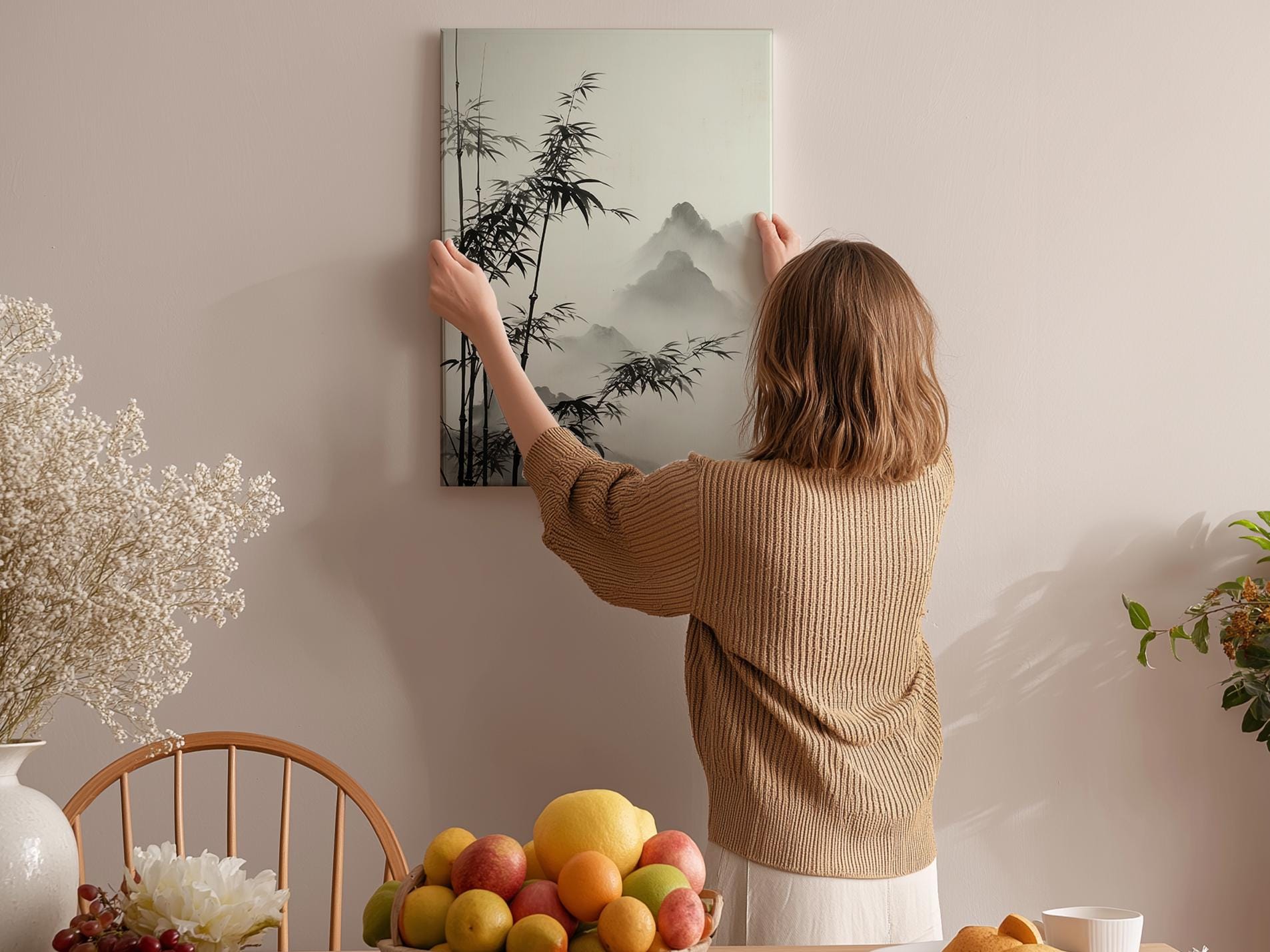 A woman hangs a black and white landscape painting on a wall in a room with a dining table, chairs, and various fruits.
