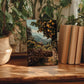 a wooden table with a framed photograph of a stone wall and a tree with oranges, surrounded by potted plants and books.