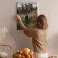 A woman hangs a framed picture of a castle on a wall in a room with a dining table, chairs, and various fruits.