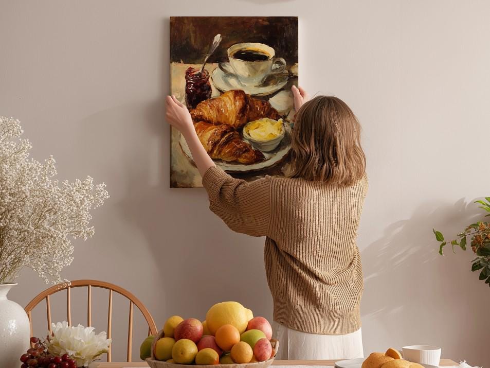 A woman hangs a painting of a breakfast scene on a wall, featuring a plate of croissants, fruit, and coffee.