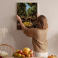 A woman hangs a framed picture of an orange grove on a wall in a room with a dining table, chairs, and various fruits.