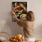 A woman hangs a framed painting of a table with various food items, including bread, fruit, and cheese, on a wall in a cozy, homely setting.