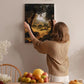 A woman hangs a framed landscape painting on a wall in a cozy room with a dining table, chairs, and various fruits.