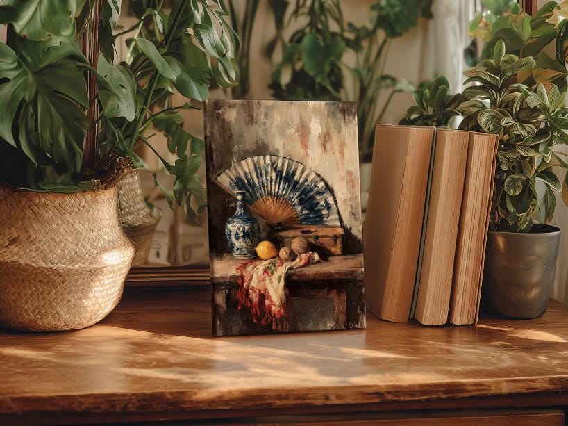 a wooden table with a framed painting of a fan and various objects, including a vase, a fruit bowl, and some books, placed on it. The table is surrounded by potted plants, creating a cozy and inviting atmosphere.