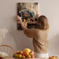 A woman hangs a painting of a still life of fruit and a fan on a wall in a room with a dining table, chairs, and various decorative items.