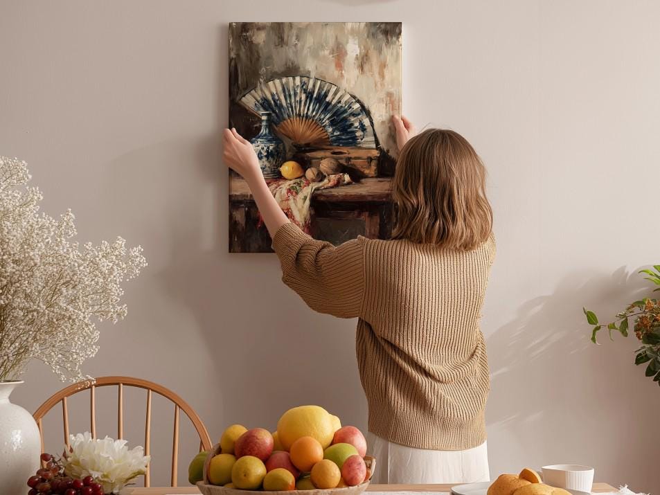 A woman hangs a painting of a still life of fruit and a fan on a wall in a room with a dining table, chairs, and various decorative items.