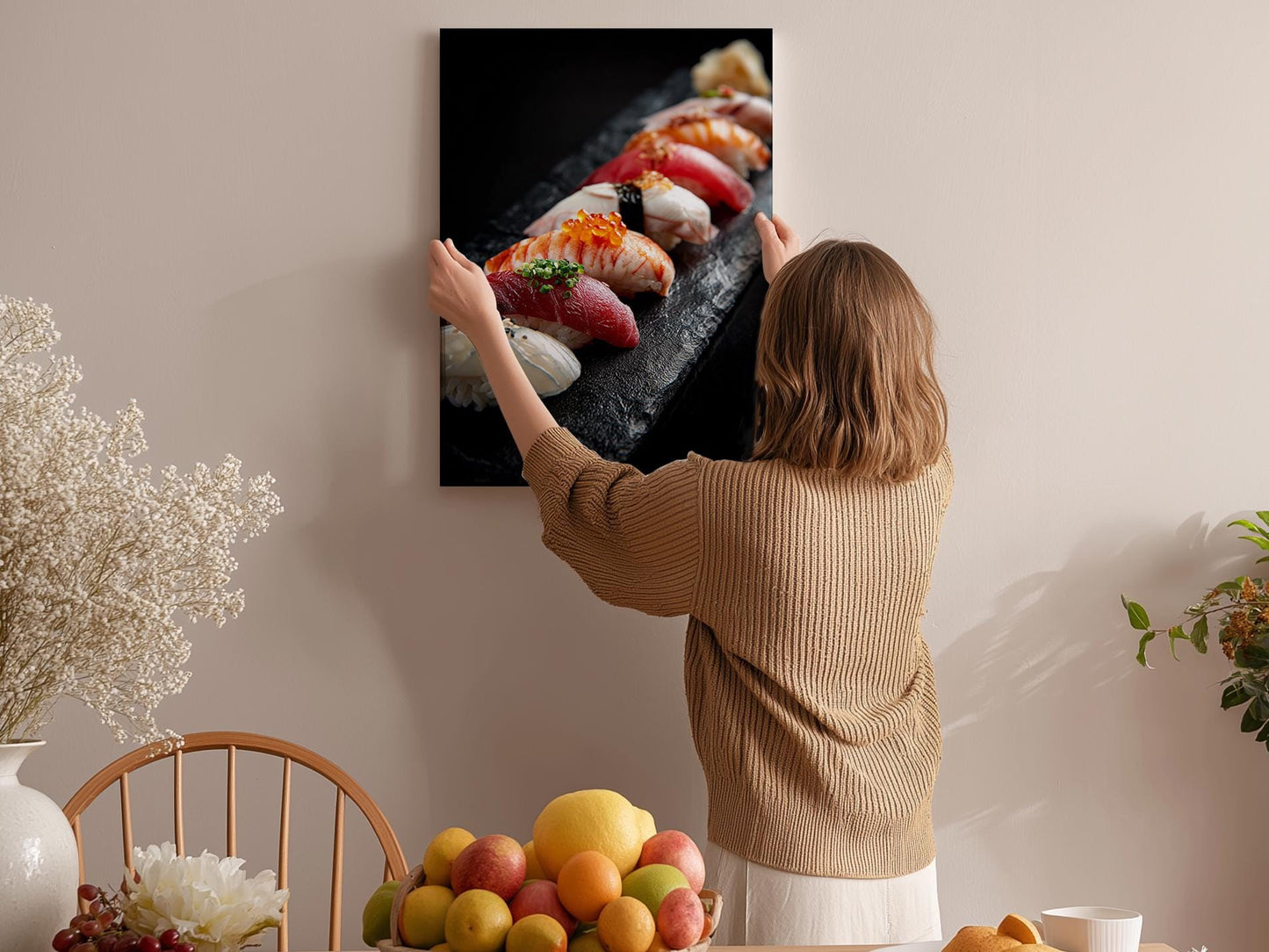 A woman hangs a framed photograph of sushi on a wall in a room with a dining table, chairs, and various fruits.