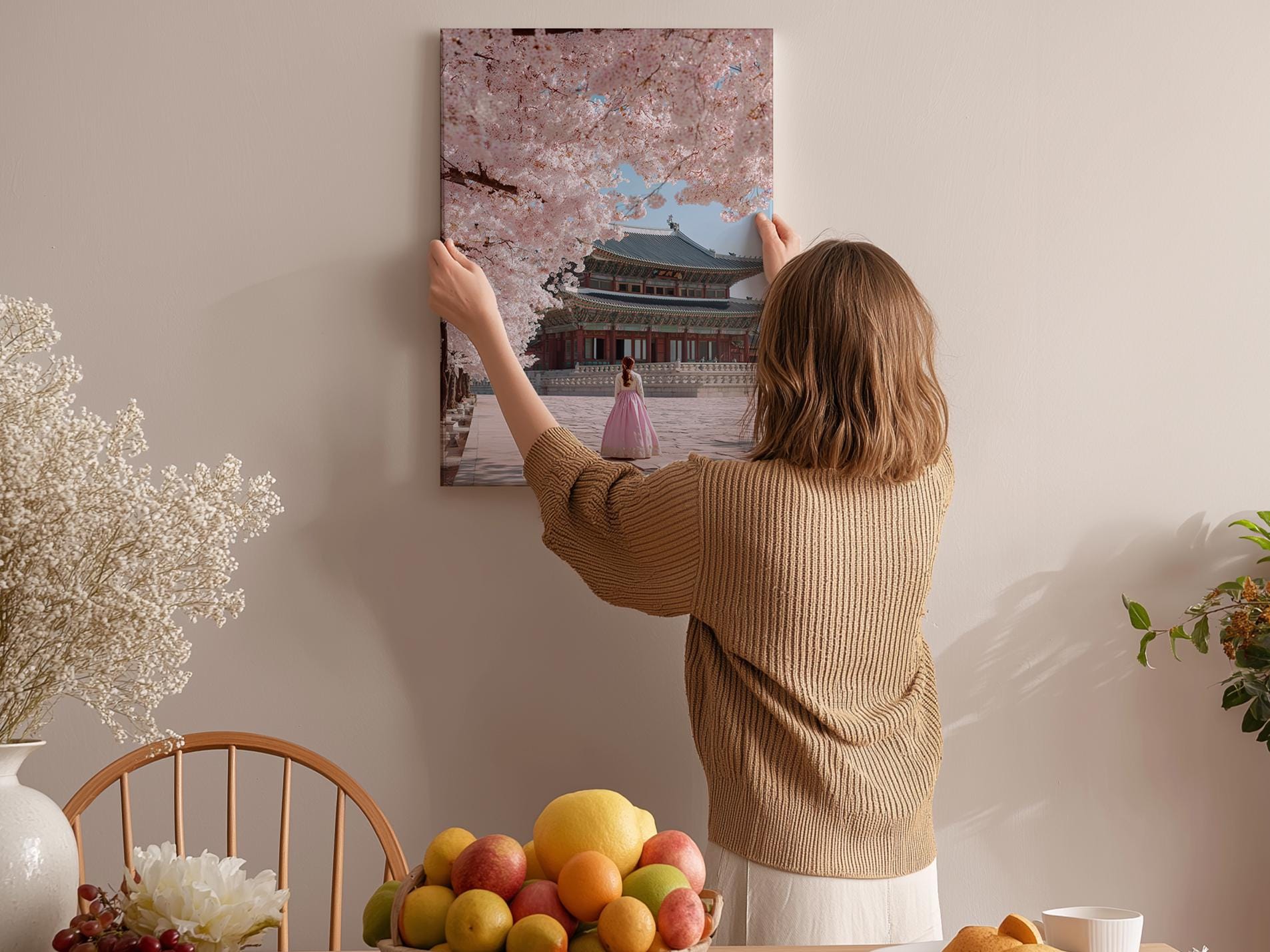 A woman hangs a framed picture of a woman in a pink dress on a wall in a room with a dining table, chairs, and various fruits.