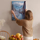 A woman hangs a framed picture of a snowy mountain landscape on a wall in a cozy room with a dining table, chairs, and various fruits.