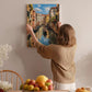 A woman hangs a framed painting of a canal scene on a wall, surrounded by a dining table with fruit and flowers.
