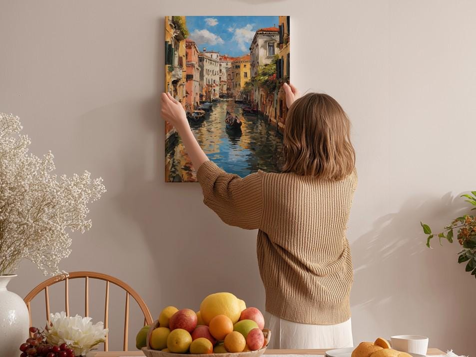 A woman hangs a framed painting of a canal scene on a wall, surrounded by a dining table with fruit and flowers.