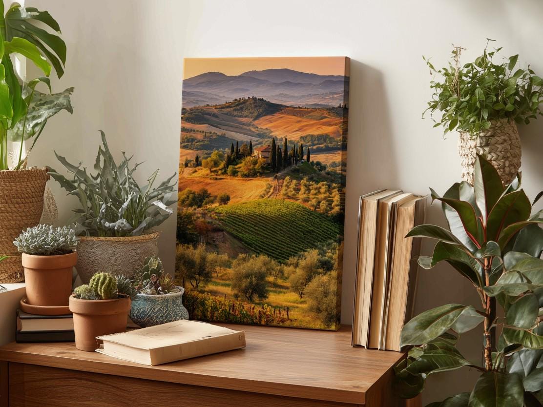 a wooden desk with a framed landscape painting depicting a scenic countryside view, surrounded by potted plants and books.