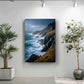 a large framed photograph of a rocky coastline with crashing waves, hanging on a white wall. To the left of the frame, there is a potted plant, and to the right, there is another potted plant.
