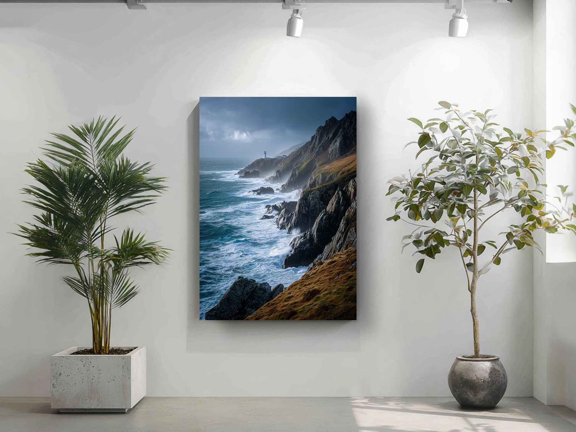 a large framed photograph of a rocky coastline with crashing waves, hanging on a white wall. To the left of the frame, there is a potted plant, and to the right, there is another potted plant.