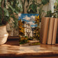a painting of a house surrounded by trees, placed on a wooden surface next to a stack of books and potted plants.