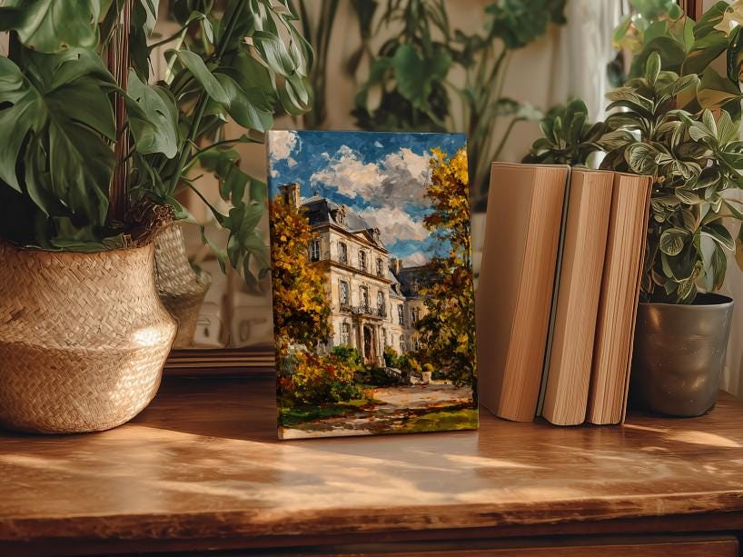 a painting of a house surrounded by trees, placed on a wooden surface next to a stack of books and potted plants.