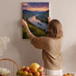 A woman hangs a framed picture of a scenic river valley on a wall in a cozy room with a dining table, chairs, and various fruits.