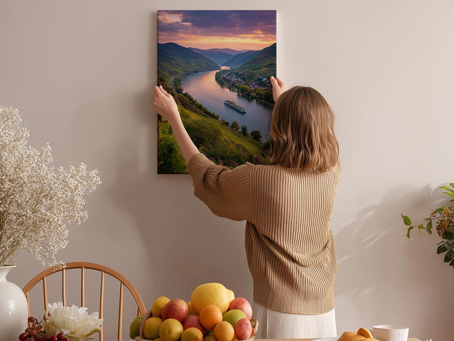 A woman hangs a framed picture of a scenic river valley on a wall in a cozy room with a dining table, chairs, and various fruits.