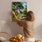 A woman hangs a painting of a house on a wall in a room with a dining table, chairs, and various fruits.