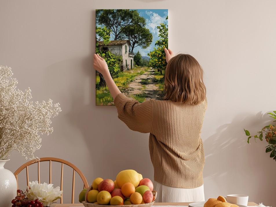 A woman hangs a painting of a house on a wall in a room with a dining table, chairs, and various fruits.