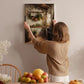 A woman hangs a painting of a kitchen scene on a wall in a cozy, homely setting.