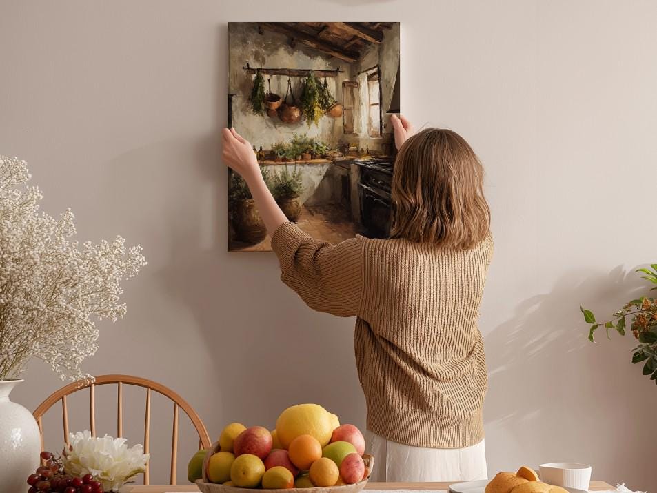 A woman hangs a painting of a kitchen scene on a wall in a cozy, homely setting.